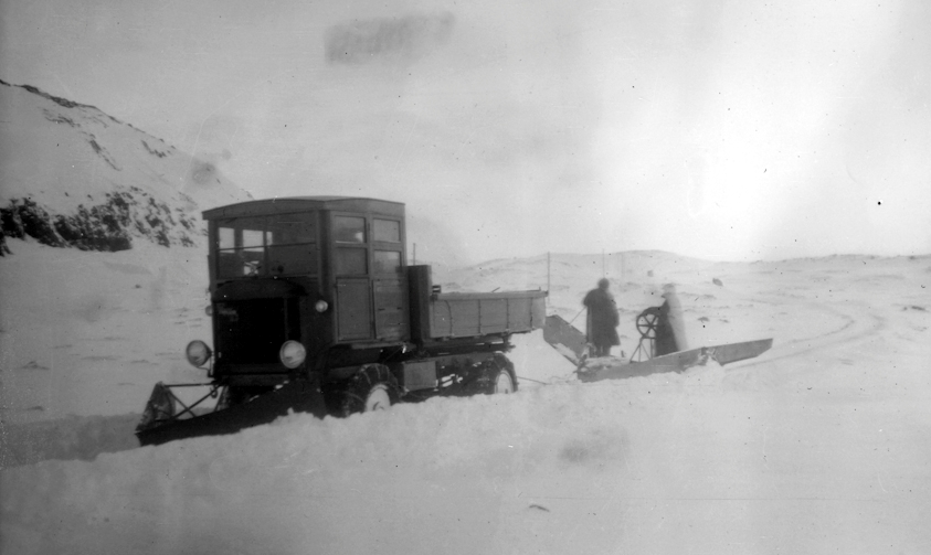 Snjómokstur í Hveradalabrekku þar sem plógur er festur aftan við bíl og minni plógur að framan. Þrjá menn þurfti að stýra tækinu. Einn ók bílnum, annar stýrði plógnum að aftan og sá þriðji sat á fremri plógnum til að þyngja hann. Mynd: Geir G. Zoëga