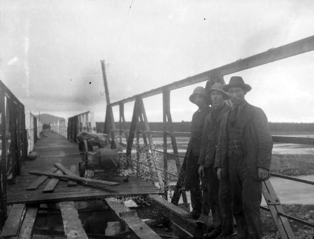 Jökulsárbrú á Sólheimasandi,1921. Brúargerð. Þrír menn standa í forgrunni myndarinnar. Fremstur gæti verið Jóhann Ólafsson sem var brúarsmiður (verkstjóri). Þar sem þeir standa úti á brúnni er ekki búið að leggja gólfið í hana svo það sést undir hana og mjög ógreinilega í hnakkann á dreng. Vinnutæki og byggingarefni sjást lengra úti á brúnni. Símastaur er á neðri hlið (hægra megin) brúarinnar sbr. mynd GZ-295. x228z A223x